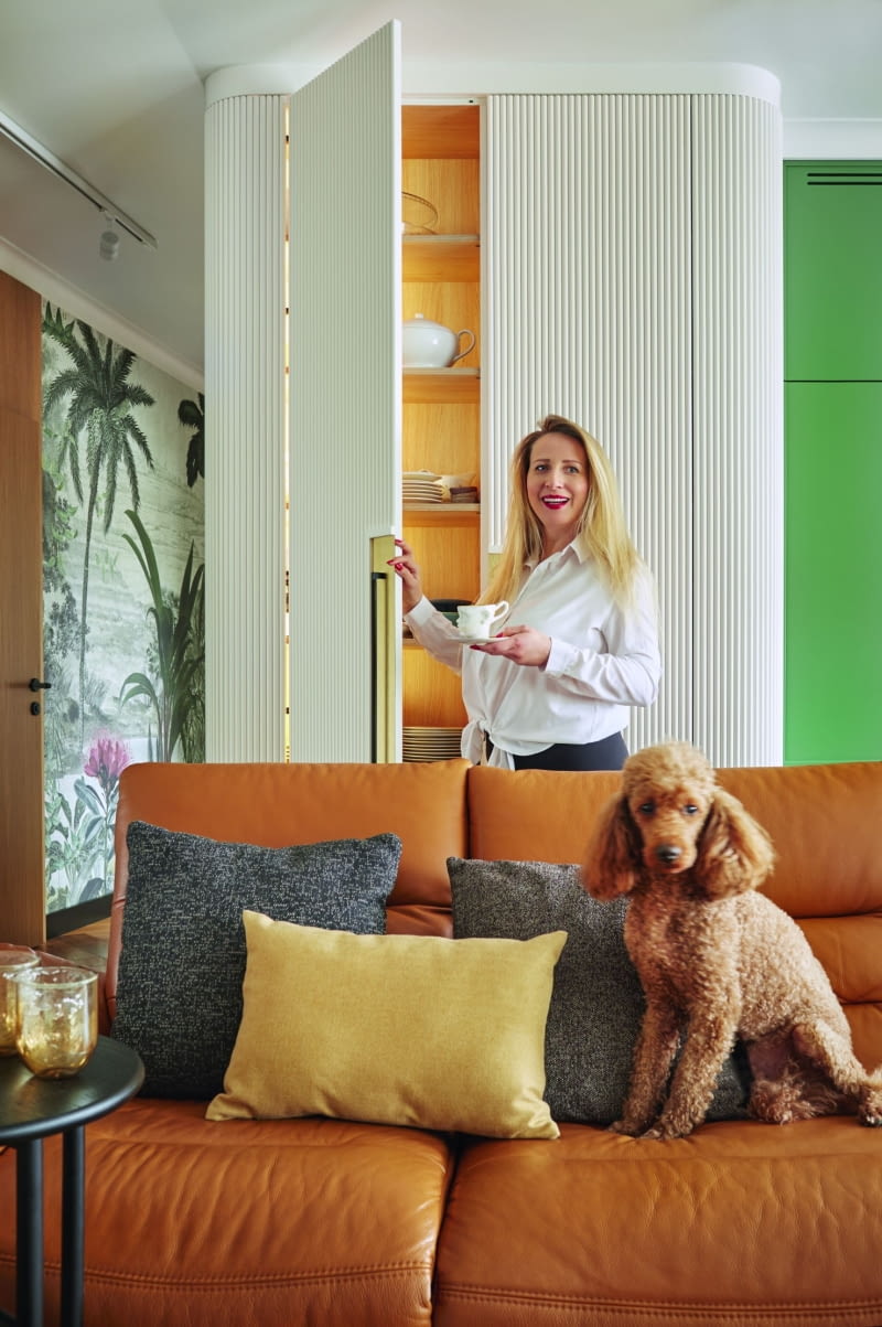 Najwspanialszym meblem kuchennym jest zdaniem gospodyni (na zdjęciu wraz z psem Choco) biała, wysoka szafa. Powstała jako uzupełniający niedużą kuchnię mebel do przechowywania wszystkiego, co nie zmieściło się w reszcie zabudowy. Wykończona pionowymi półwałkami szafa w środku wyłożona jest drewnianym fornirem. Jest też podświetlana. W takiej scenerii pięknie prezentuje się porcelanowa zastawa stołowa. To nie jedyny motyw obłości, który wykorzystała w aranżacji wnętrza architektka Magdalena Chrzanowska. Krągłości są obecne w tapicerowanej ściance przy wejściu, w zabudowie kuchennej i delikatnych wałeczkach na frontach mebli. Obły, bo okrągły jest też stół oraz forma stojących przy nim krzeseł. Jadalniany mebel wyszukała pani domu. Jest pięknie wykończony fornirem pasującym do koloru parkietu i ma czarny, szklany blat, wpisujący się w zamysł czarnych dodatków w tym kolorze, na przykład krzeseł w czarno-białą pepitkę.