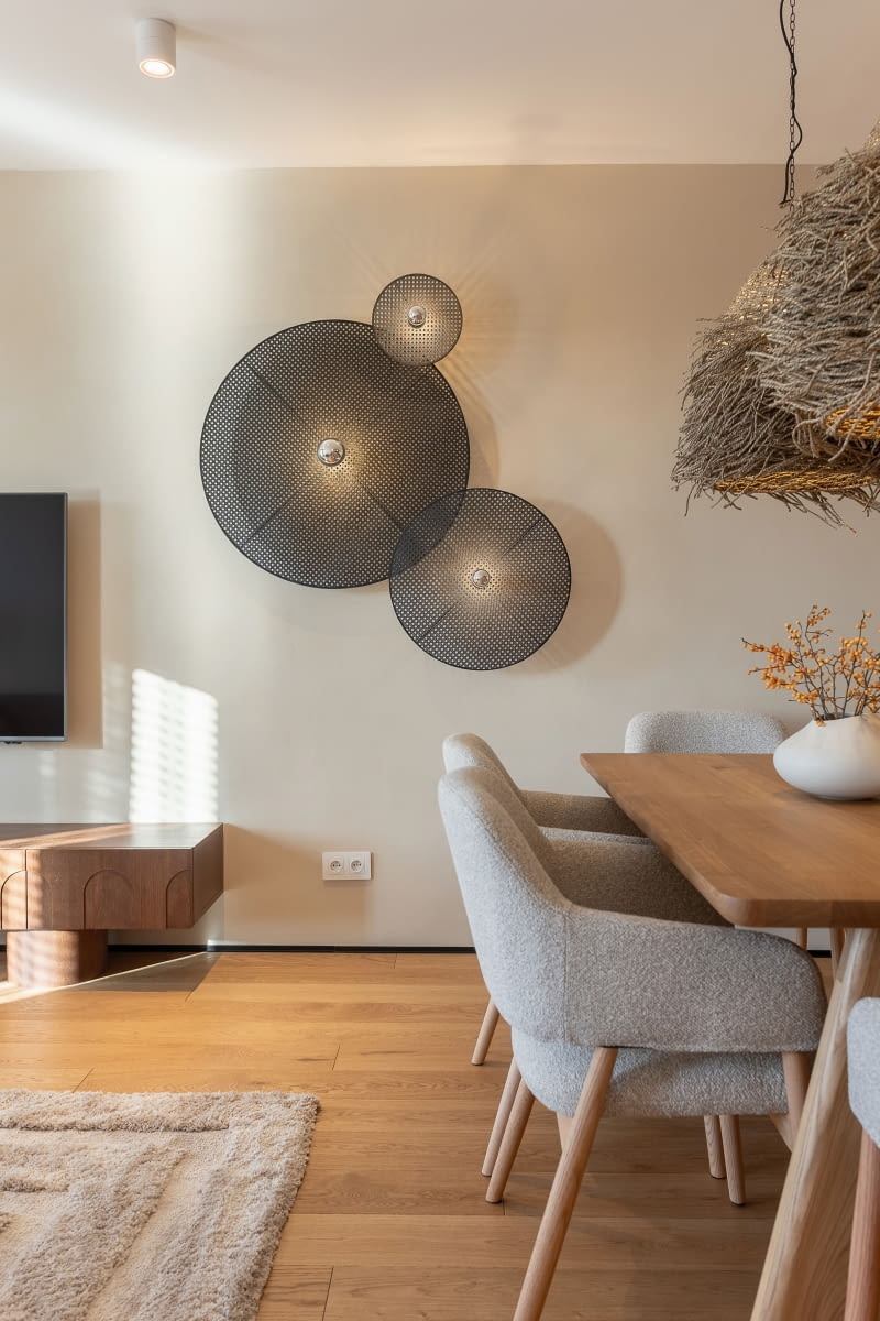 Na ścianie obok telewizora wiszą trzy czarne, ażurowe kinkiety z rattanu marki Aromas del Campo, dostarczone przez studio sztuka-swiatla.pl. Przy dębowym, eleganckim stole od Smreki stanęły tapicerowane krzesła z lectus24.pl.