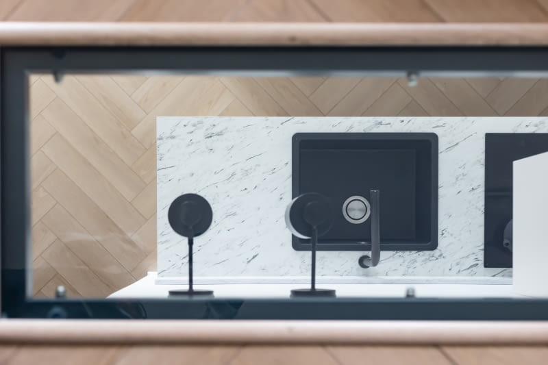 Pomiędzy piętrami, w suficie, nad aneksem kuchennym deweloper przewidział otwór. Ciekawy detal architektoniczny zyskał oprawę ze szkła w antracytowej, podobnie jak konstrukcja i balustrada schodów, stalowej ramie. Przeszklenie doświetla kuchnię i dodaje dodatkowej lekkości całej przestrzeni.