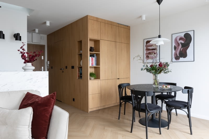 Ściana i zabudowa meblowa we wspólnym wykończeniu drewnem tworzą atrakcyjną, zwartą, architektoniczną formę – swego rodzaju kubik. Skrywa on łazienkę, lodówkę oraz szafę do przechowywania.