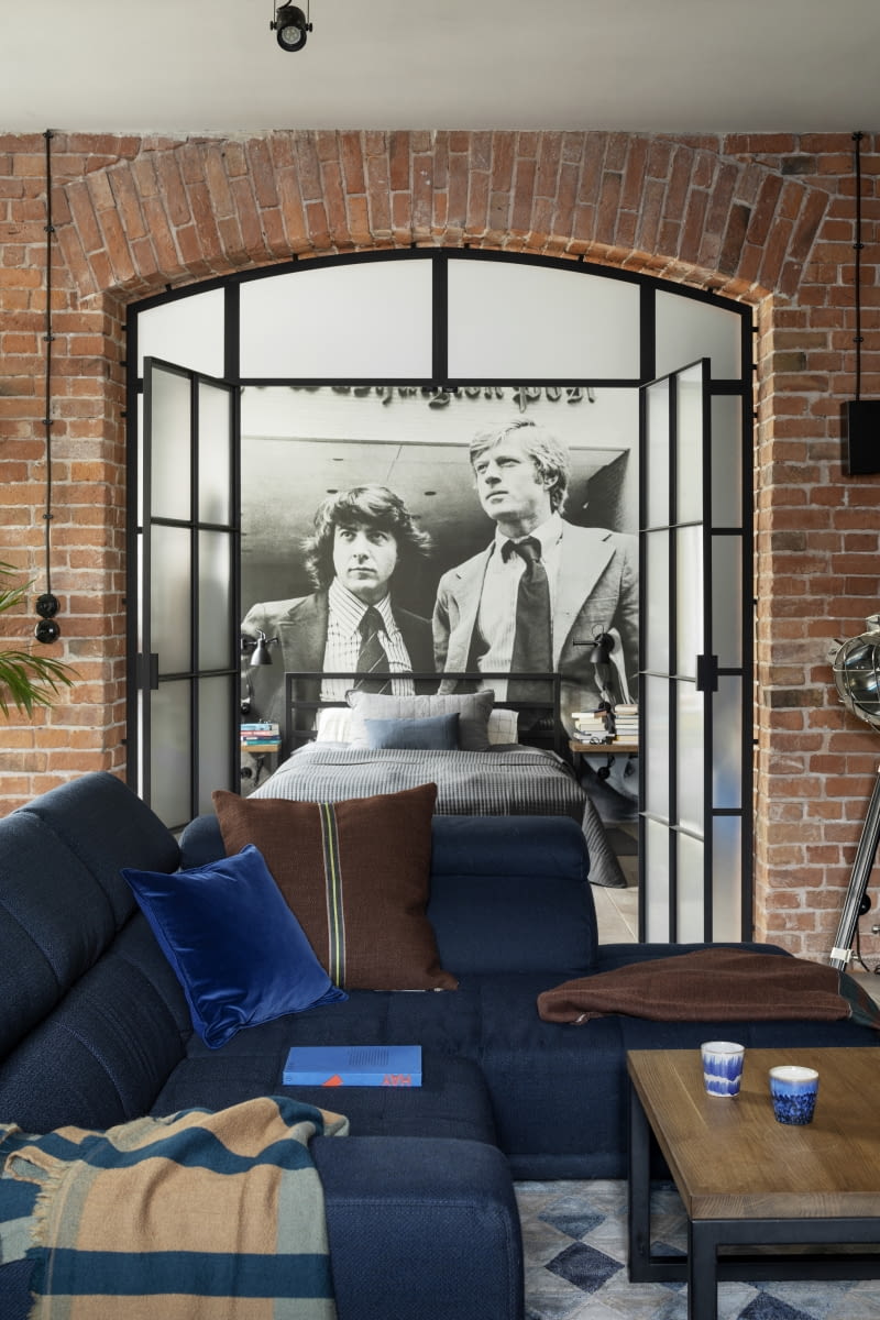 Dominujące w aranżacji kolory naturalnej cegły, drewna, betonu, bieli, ciemnej szarości i czerni – typowych dla industrialnych wnętrz – ożywiają granatowe, tapicerowane meble. Ładnie komponują się z nimi tkaniny w barwach brązu. Dekoracyjne pledy i poduszki ułożone na kanapie pochodzą ze sklepu NAP, a kubki w odcieniu sofy są marki HKliving.