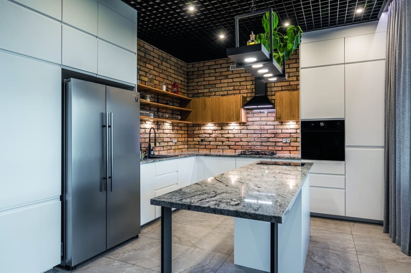 W przylegającej do salonu kuchni centralne miejsce zajmuje wyspa z granitowym blatem, specjalnie zamówionym w zakładzie kamieniarskim.  Na podłodze leżą włoskie płytki firmy Imola Ceramica, a ścianę w głębi efektownie pokryto ćwiartkami cegieł. Kratownica u góry to sufit rastrowy,  często spotykany w sklepach czy magazynach – dobrze wpisuje się w industrialny charakter wnętrz