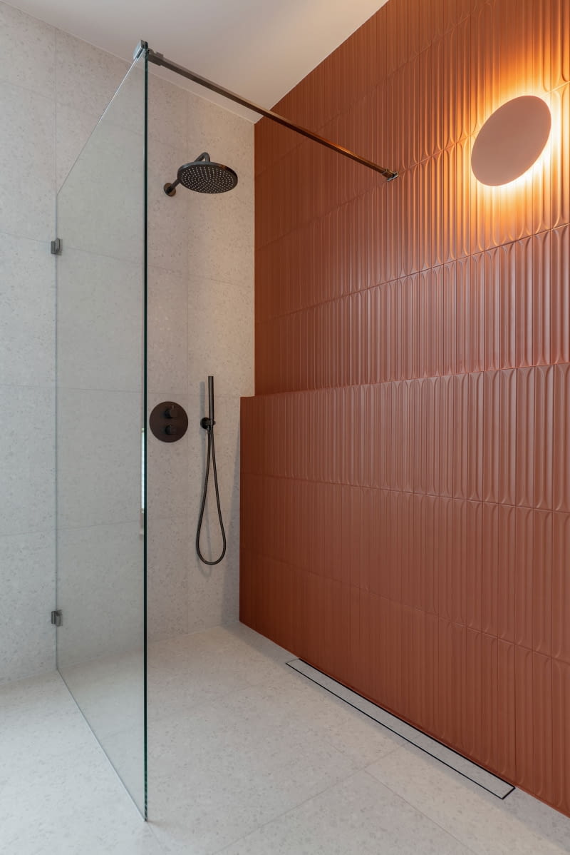 Strukturalne, ceglaste płytki  Equipe wprowadzają do łazienki  wyrazisty akcent dekoracyjny. W strefie prysznica pokrywają całą ścianę, a ich rytm przełamuje delikatnie wysunięta przedścianka tworząca uskok-półkę na kosmetyki. Trójwymiarowy efekt płytek uwydatnia punktowe światło okrągłego kinkietu marki Nowodvorski.