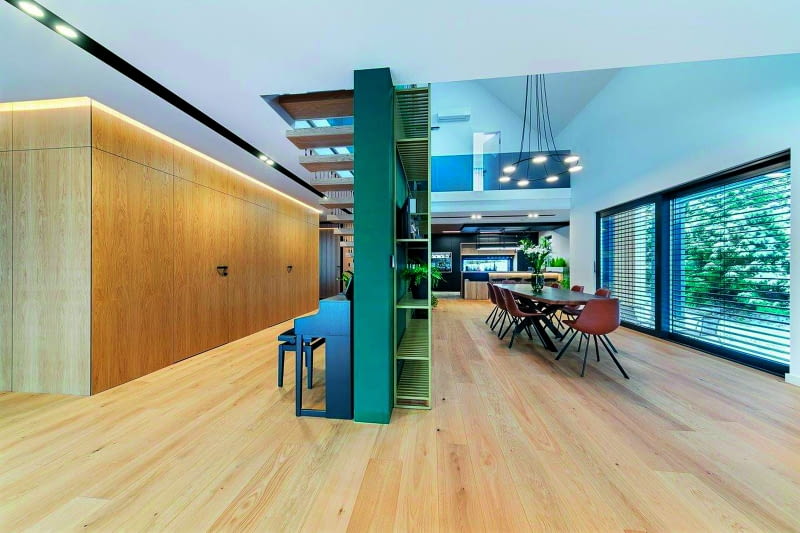 Zwykła, luźno stojąca ściana działowa o długości ok. 5 m potrafi zamienić otwartą przestrzeń dzienną w strefy o odmiennym charakterze – jeśli ją tylko odpowiednio doposażymy.  Ażurowy regał po prawej wnosi fantazję do jadalni, a po lewej górą czarują lekkością schody,  zaś pod nimi przewidziano miejsce na uniesienia pianisty