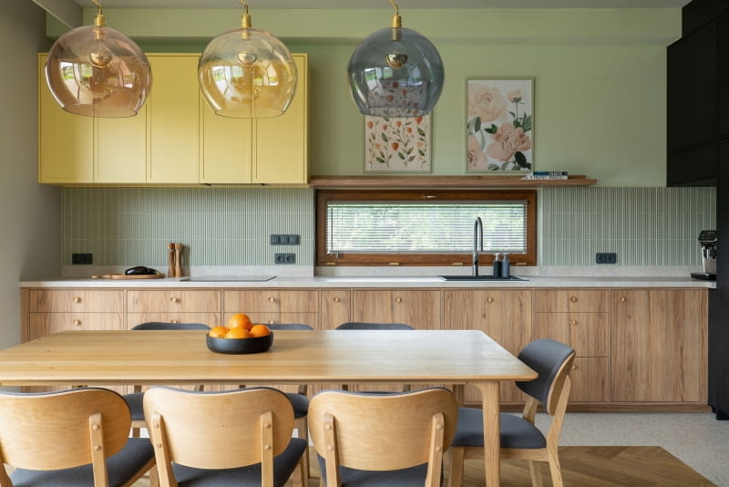 Długi, prostokątny stół do rodzinnych spotkań powtarza podłużny układ kuchni. Drewniany mebel pochodzi z Westwing, krzesła  od polskiej marki Woodica, a szklane lampy  z EBB & Flow.