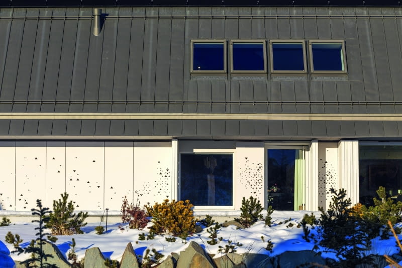 Widok na fragment jednej z bocznych elewacji – konkretnie tę z głównym wejściem do budynku. Cały parter nakryty jest dwukondygnacyjnym skośnym dachem z ciemnej blachy cynkowo-tytanowej Widok na fragment jednej z bocznych elewacji – konkretnie tę z głównym wejściem do budynku. Cały parter nakryty jest dwukondygnacyjnym skośnym dachem z ciemnej blachy cynkowo-tytanowej