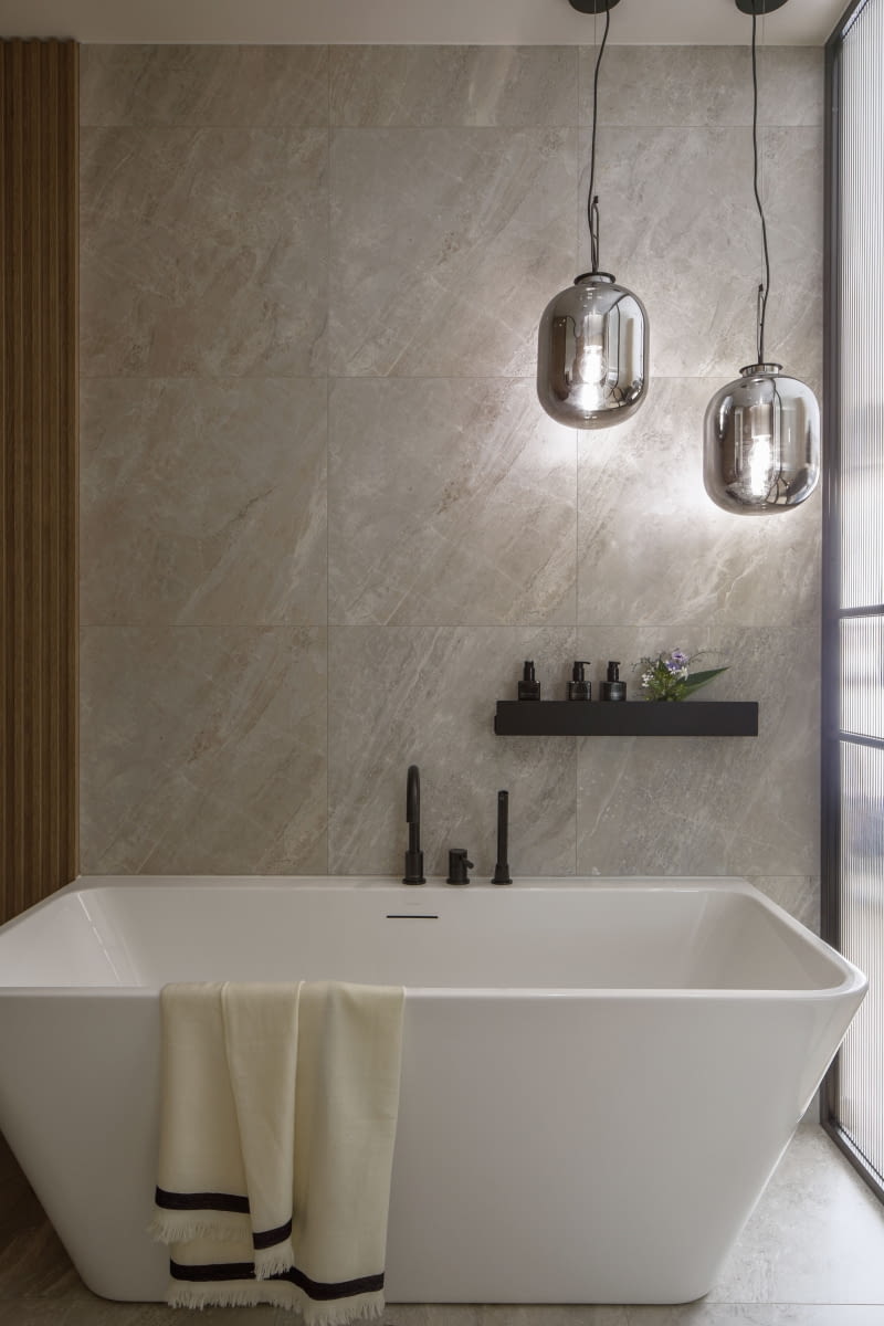 Światło dwóch wiszących lamp Maxlight Honey podkreśla rysunek kamienia odwzorowanego na gresie Porcelanosa Indic, którym zabezpieczono ścianę przy wannie marki Massi. Czarna armatura pochodzi z oferty firmy Kohlman.