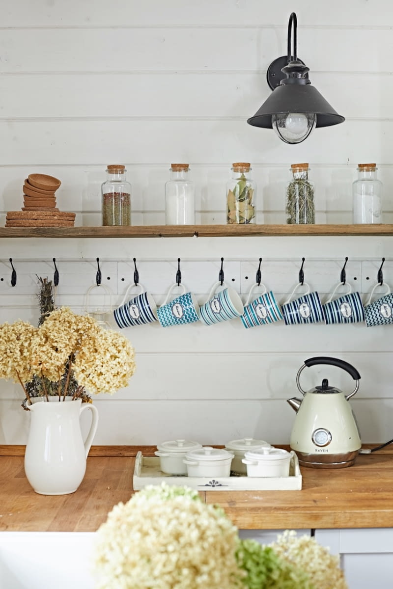 Klimatyczne akcenty w postaci rustykalnej półki z haczykami na kubki oraz czarnego kinkietu czy dobranej z tej samej serii lampy wiszącej uzupełniają wystrój kuchni.