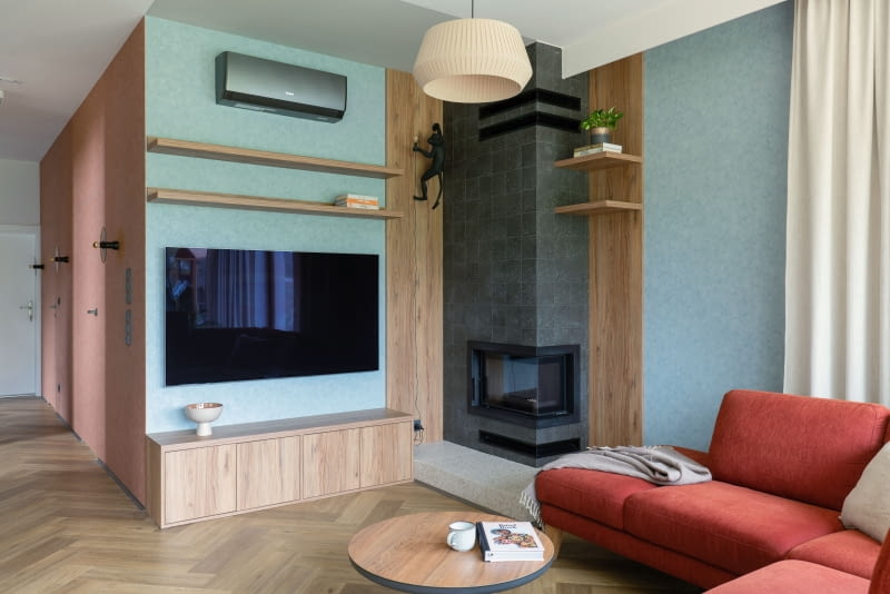 W salonie, podobnie jak w całym domu, ciepłe kolory przeplatają się z zimnymi. Tutaj odcienie bordo, beżu i drewna kontra błękit przełamuje mocny, graficzny akcent w postaci obudowy kominka  z niemal czarnych płytek WOW z kolekcji Stardust Pebbles Nero  - ich powierzchnia przypomina ręcznie wyrabianą glinę.