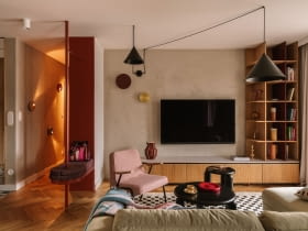 Salon wyposażono w welurową, jasnozieloną sofę z Etap Sofa, różowy fotel Chierowskiego marki 366 Concept oraz czarne stoliki kawowe Tamo. Podłogę zdobi dywan ze sklepu Rugvista. Strefę doświetlają wiszące nad kompozycją mebli czarne lampy od Kolorowe Kable i dwa kinkiety od ENOstudio. Ścianę z telewizorem wykończono strukturalnym tynkiem w beżowym odcieniu.