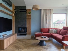 . Obok kominka stanęła duża, rodzinna sofa w bordowym odcieniu oraz stolik łączący drewniane i czarne elementy – zestaw spina całą strefę wypoczynkową.