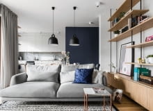 We wnętrzu ważną funkcję pełni kolorystyczny kontrast między granatową bryłą a wyposażeniem wnętrz w białych i szarych barwach, z dodatkiem naturalnego drewna. Ożywia przestrzeń. Salon otwarty na kuchnię. Aranżacja w kolorach granatu, szarości, bieli, z dodatkiem drewna