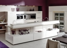 1. WYPRACOWANA ELEGANCJA. Ustawiona na tle wrzosowej ściany zabudowa z kremowej płyty MDF (matowej i lakierowanej na wysoki połysk) wygląda raczej jak część reprezentacyjnego wnętrza niż miejsce do przygotowywania posiłków. Wrażenie to jest tym mocniejsze, że przykrywający wyspę blat roboczy z płytą grzejną i zlewozmywakiem jest dyskretnie zasłonięty niewysoką nadstawką, dzięki czemu osoby siedzące przy stole nie widzą kuchennego rozgardiaszu. W wyspie od strony jadalni znajdują się pojemne szuflady (np. na sztućce i obrusy) i długa otwarta półka (może pełnić funkcję tzw. pomocnika).