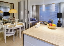 Tłem dla zabudowy kuchennej jest powierzchnia pomalowana na powtarzający się w całym mieszkaniu jasnoszary kolor. Zabieg ten ożywia biel szafek, które delikatnie nabierają odcienia ściany. Wiszące nad barkiem lampy (Leroy Merlin) wyznaczają przestrzenną granicę kuchni. Aranżacja wnętrz