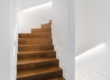 Betonowe schody zabiegowe prowadzące na piętro zostały obłożone drewnem. Zastosowanie takiego materiału w ciemnym kolorze w połączeniu z bielą ścian sprawia, że ten fragment wnętrza jest elegancki i nowoczesny zarazem Betonowe schody zabiegowe prowadzące na piętro zostały obłożone drewnem. Zastosowanie takiego materiału w ciemnym kolorze w połączeniu z bielą ścian sprawia, że ten fragment wnętrza jest elegancki i nowoczesny zarazem