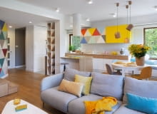 Architekci stworzyli przestronną, jasną i przytulną przestrzeń, którą wypełniają ciepłe kolory, proste formy mebli oraz naturalne materiały z drewnem na czele. Aranżacja kolorowego salonu. Motyw przewodni aranżacji wnętrza kolorowe trójkąty