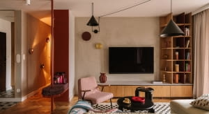 Salon wyposażono w welurową, jasnozieloną sofę z Etap Sofa, różowy fotel Chierowskiego marki 366 Concept oraz czarne stoliki kawowe Tamo. Podłogę zdobi dywan ze sklepu Rugvista. Strefę doświetlają wiszące nad kompozycją mebli czarne lampy od Kolorowe Kable i dwa kinkiety od ENOstudio. Ścianę z telewizorem wykończono strukturalnym tynkiem w beżowym odcieniu.