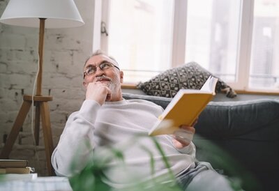 Ältere Person sitzt mit einem Buch auf einem Sofa in einem Wohnzimmer.