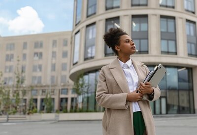 Person steht mit Tablet und Unterlagen vor einem modernen Bürogebäude.