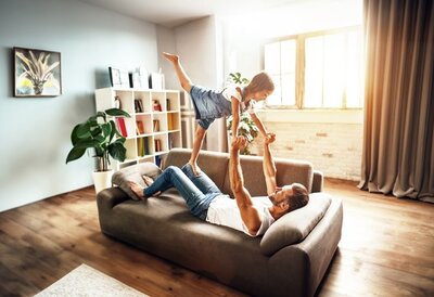 Erwachsene Person hebt ein Kind auf einem Sofa in die Luft.