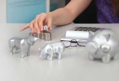 Münzstapel und Sparschweinfiguren liegen neben Taschenrechner und Brille auf einem Tisch.