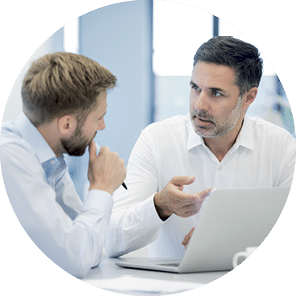 Two adult Caucasian businessmen in open neck shirts discussing business project around a laptop computer in a modern office 