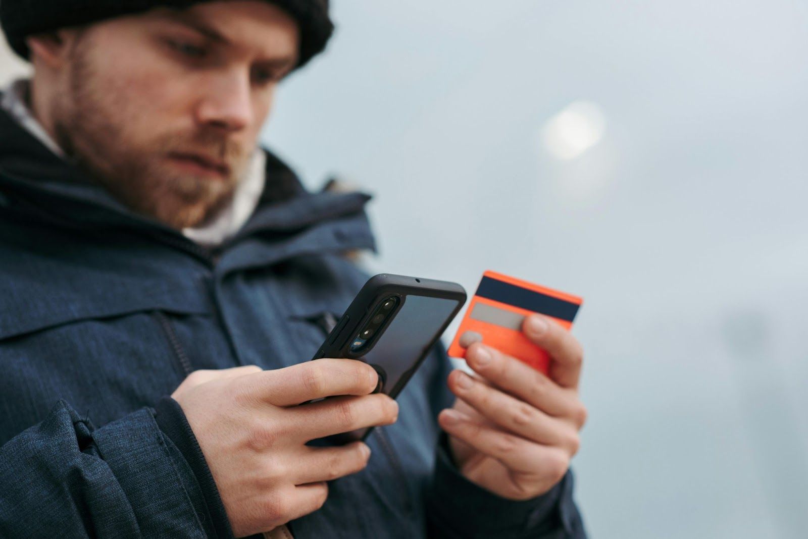 Ein Mann hält ein Smartphone in der einen und eine orangefarbene Kreditkarte in der anderen Hand. 