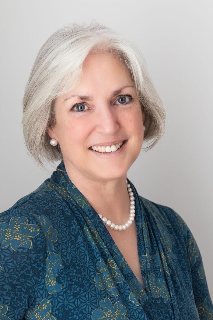 Sara Price Celebrant, a white lady with white hair, wearing a blue patterned dress with a pearl necklace and earrings.