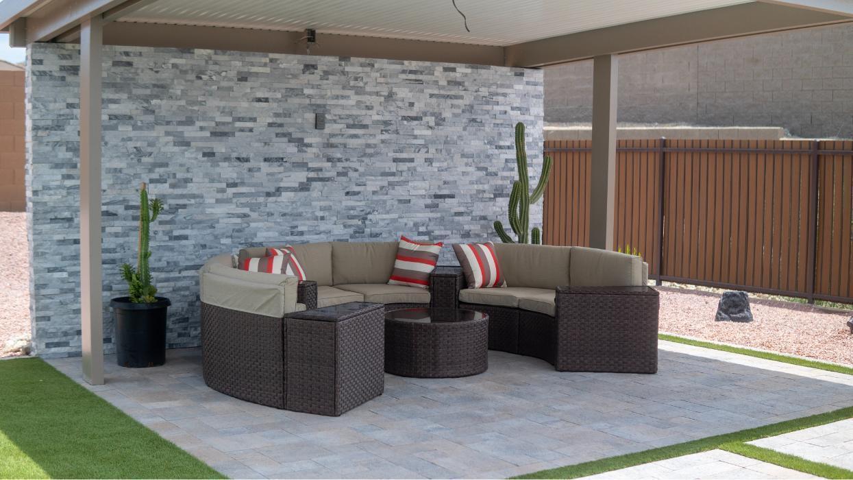 Covered patio with stone accent wall and outdoor furniture. AE Outdoor Living&rsquo;s hardscape and shade structures add usable backyard space.