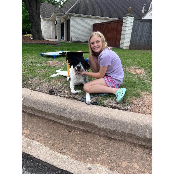 A girl sitting on a curb hugging a black-and-white dog with a house and yard in the background.