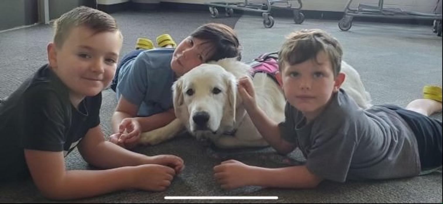 Three kids are lying on the floor, cuddling with a white dog between them.