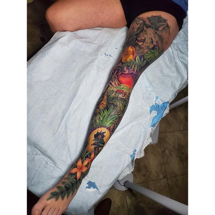 Jurassic Park themed leg sleeve tattoo. Full color realism featuring dinosaurs, tropical plants, and iconic movie details.