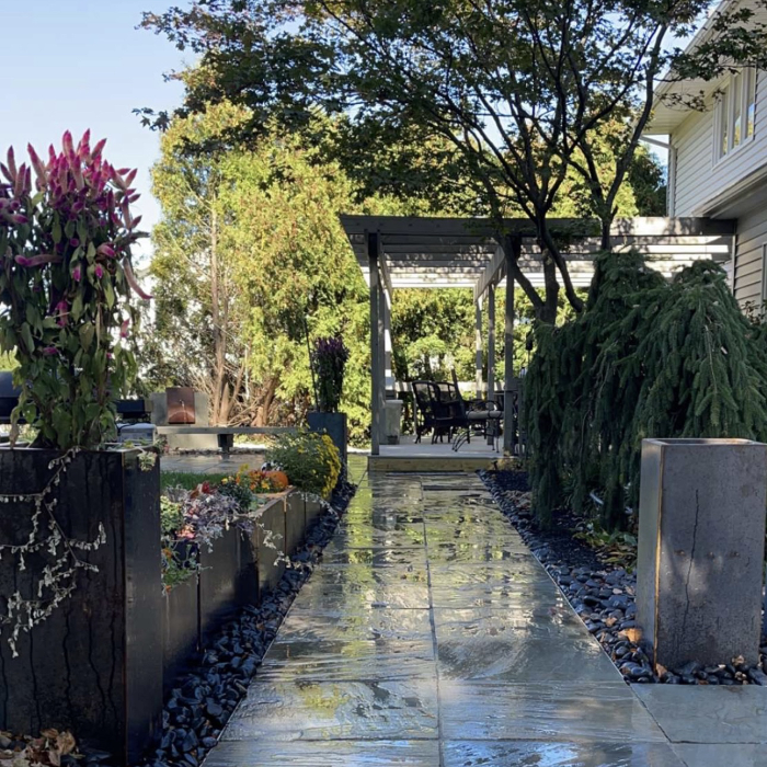 Elevate your curb appeal with hardscaping that makes a statement. This serene walkway combines wet-look stone pavers, decorative black river rock, and modern planters to create a textured, inviting path leading to a cozy pergola retreat.