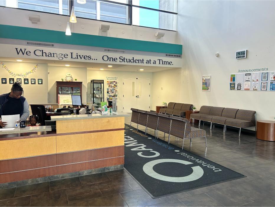 The welcoming lobby and reception area at Camen Behavioral Services, featuring the motivational slogan "We Change Lives... One Student at a Time," comfortable seating for families, and a community resources station.