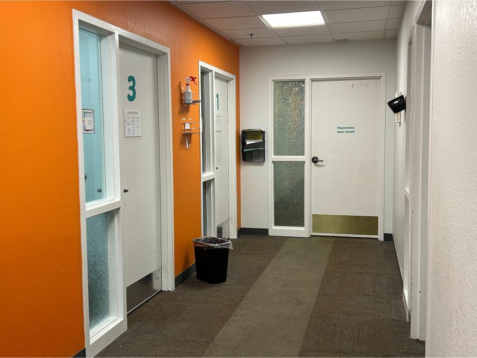 A clean, brightly lit hallway with an orange accent wall leading to numbered private therapy rooms at Camen Behavioral Services.
