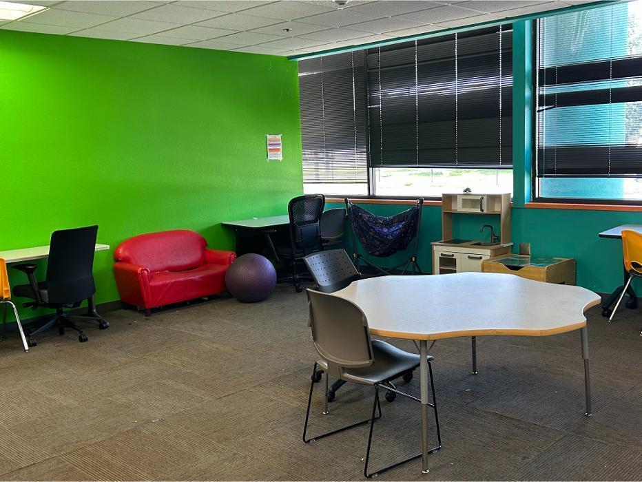 A vibrant therapy room at Camen Academy with a lime green accent wall, featuring a red child-sized sofa, a sensory swing, a play kitchen, and individual workstations.