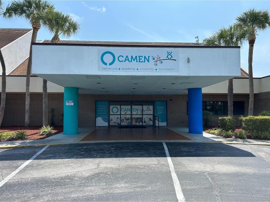 Close-up of the main entrance at Camen Behavioral Services, showing the company logo on the overhang, teal and blue support pillars, and professional window decals detailing services like Behavior Analysis and Social Skills Programs.