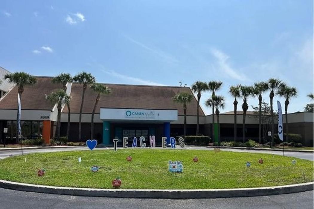 Wide shot of the Camen Behavioral Services building exterior featuring a circular green lawn with "TEACHERS" lawn signs and palm trees under a clear blue sky.