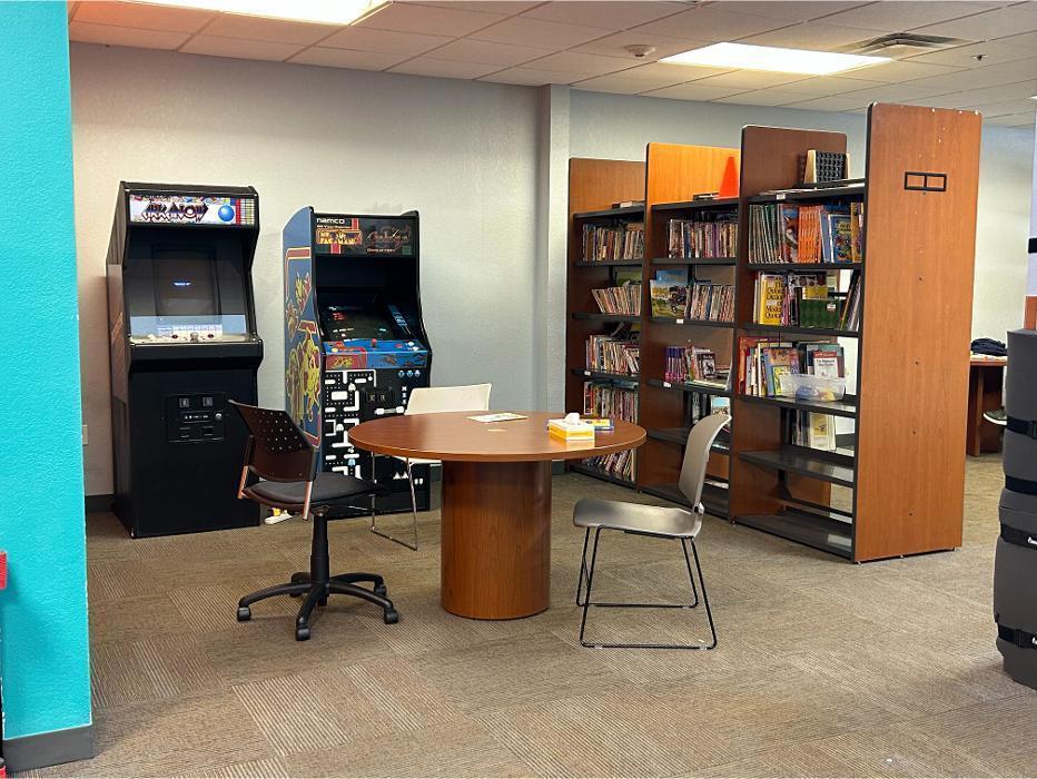 A student-focused area at Camen Academy combining learning and play, featuring tall wooden bookshelves filled with books, a round study table, and classic arcade machines like Ms. Pac-Man/Galaga for reinforcement and recreation.