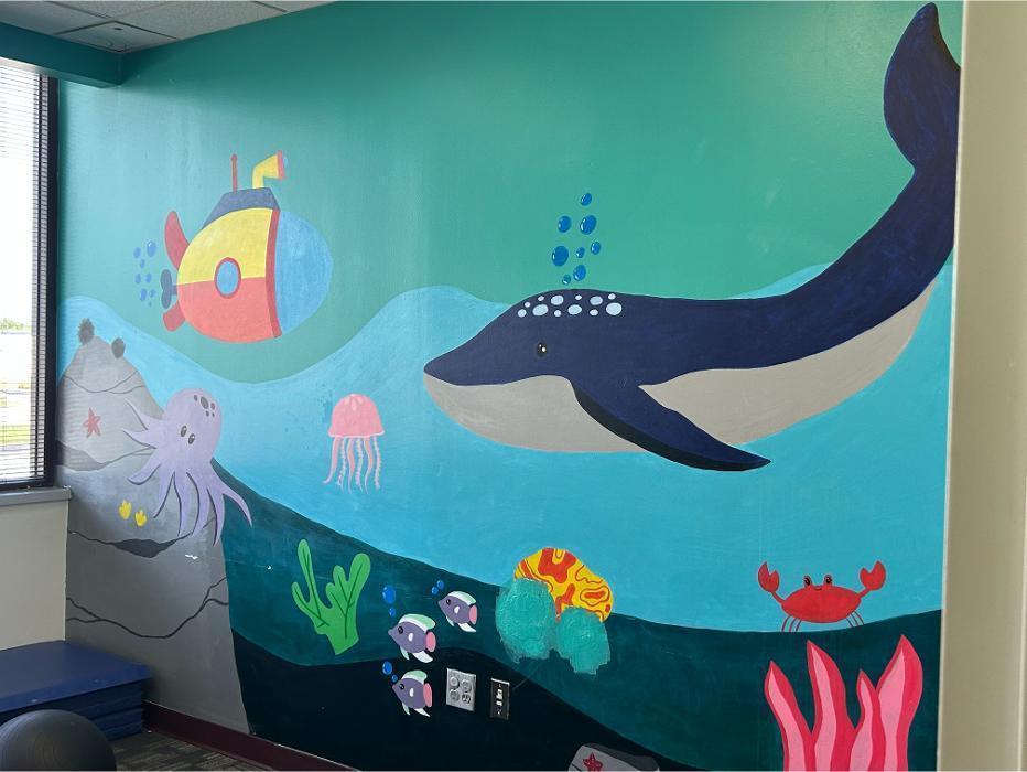 A large, colorful underwater-themed mural on a therapy room wall featuring a whale, submarine, octopus, and crab, designed to create a welcoming environment for children.