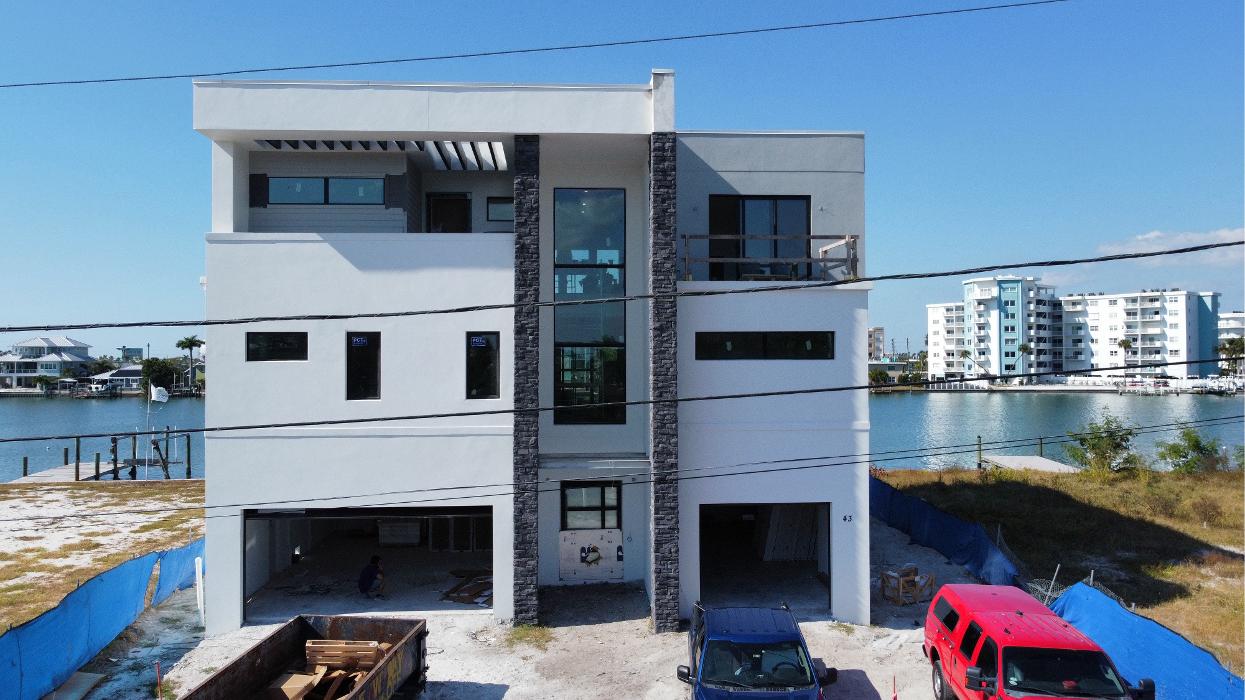 Front exterior rendering of a modern multi-story waterfront home with large windows, balconies, rooftop space, parked cars, and water views on both sides.