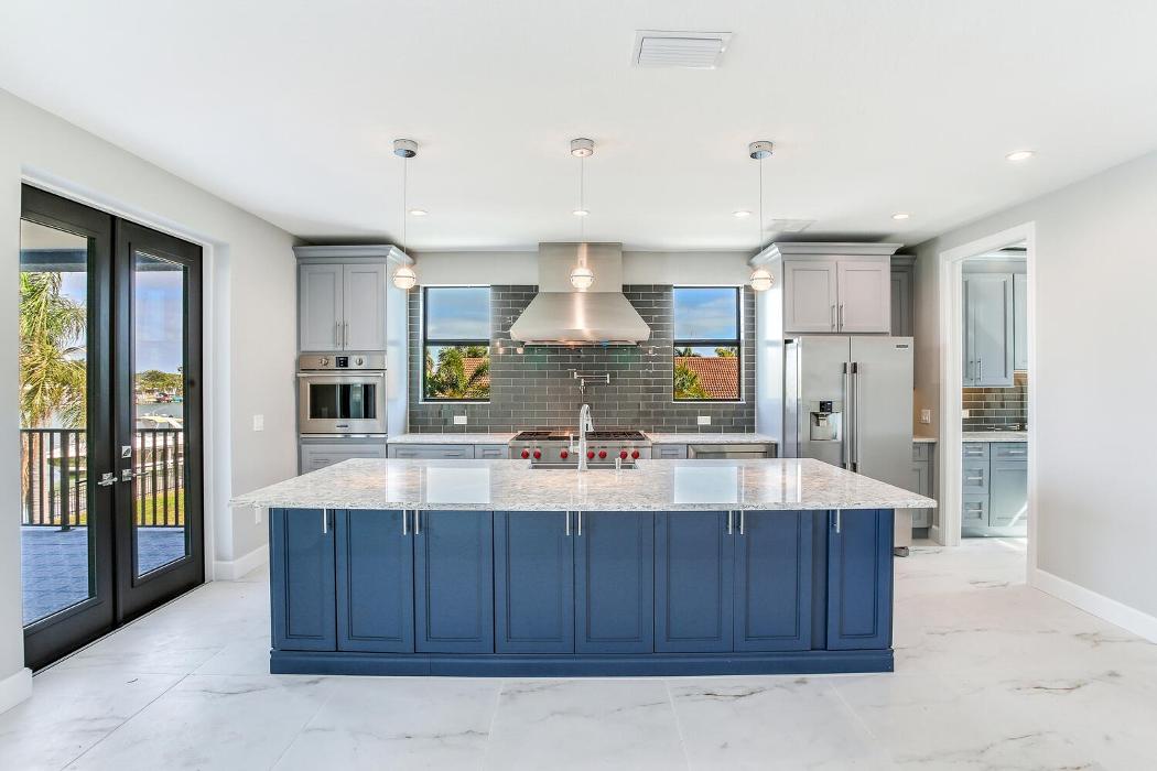 Front view of a contemporary kitchen featuring a large blue island, pendant lighting, stainless steel range hood, and wide open surrounding space.