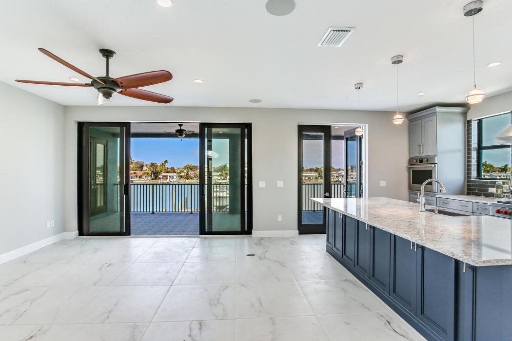 Bright open-concept kitchen and living area with light tile flooring, blue lower cabinets, a large island, ceiling fan, and sliding glass doors.