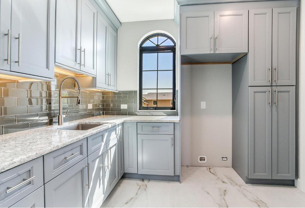 Contemporary kitchen featuring gray cabinetry, a stainless steel refrigerator, light countertops, and a decorative arched window above the sink.