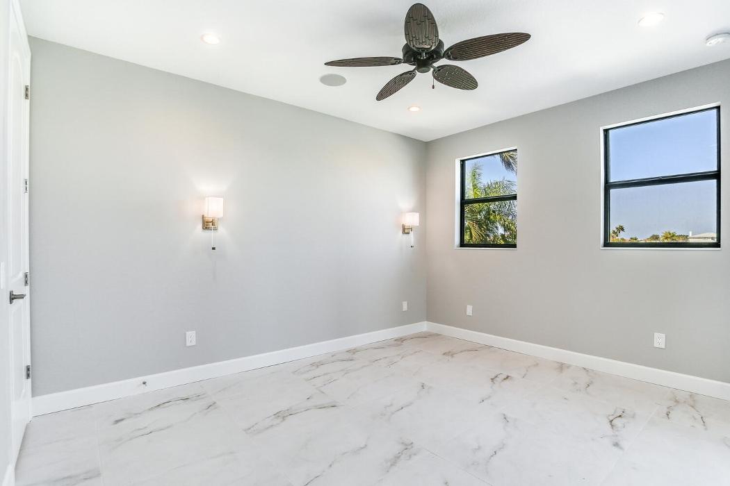 Spacious bedroom with light tile flooring, gray walls, a ceiling fan, wall sconces, and wide windows bringing in natural light.