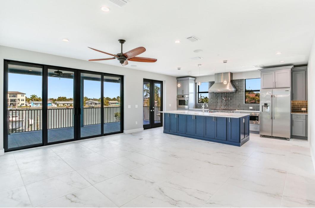 Wide view of an open-concept main floor with large sliding glass doors, light tile flooring, ceiling fans, and a modern kitchen with blue cabinets and an island.