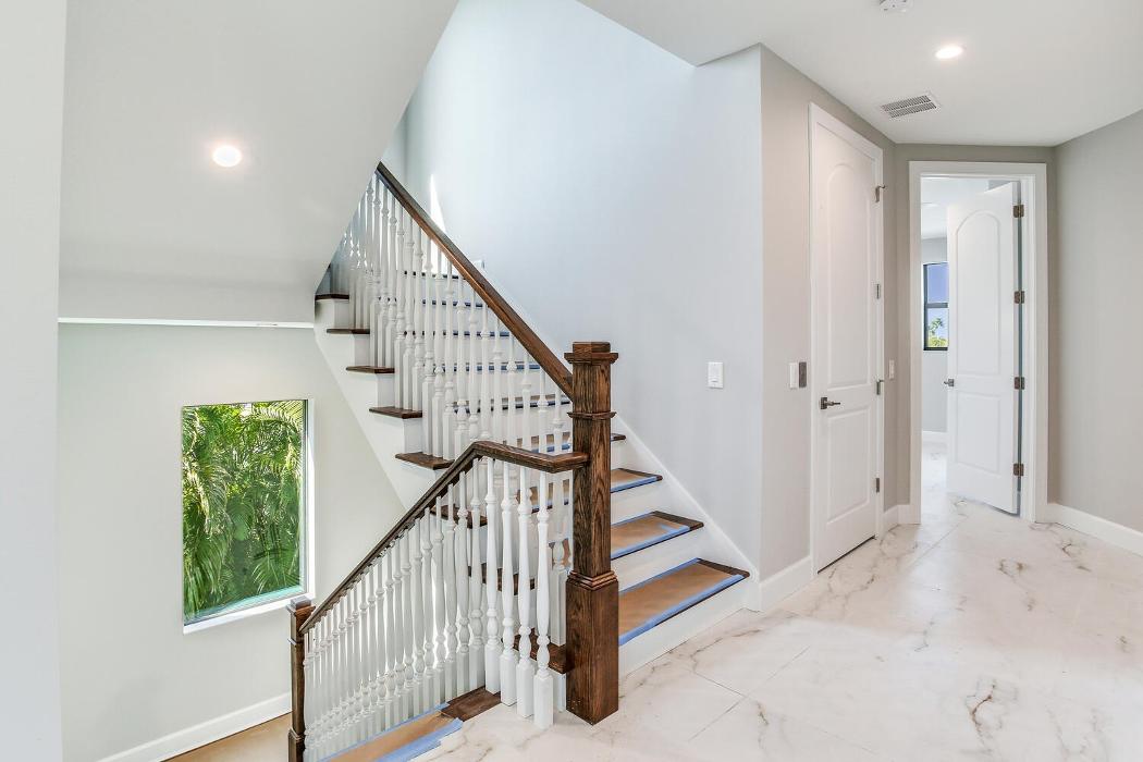 Modern stairwell and hallway with light tile floors, a wood-and-metal staircase railing, white walls, and natural light from nearby windows.