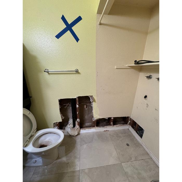 An unfinished bathroom or utility room with a section of drywall removed near the bottom of the wall and blue tape marking an area above.