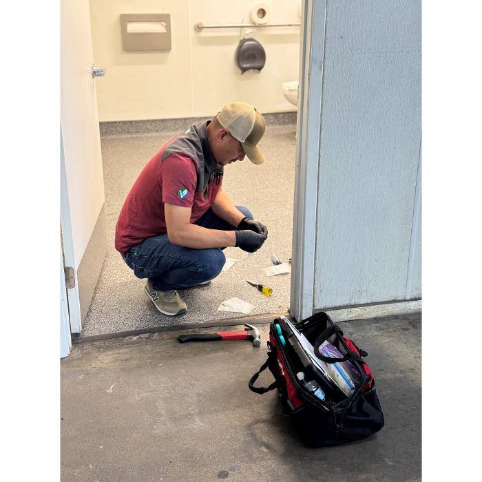 A worker kneeling near a wall with a tool bag beside him, appearing to inspect or perform work at floor level.
