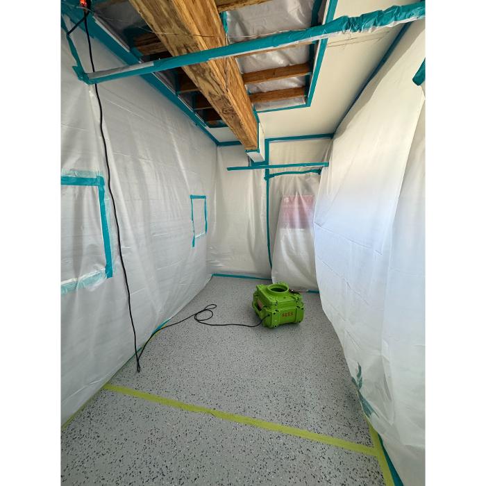 A long enclosed area or basement section lined with white plastic sheeting and blue tape, with exposed ceiling beams overhead.